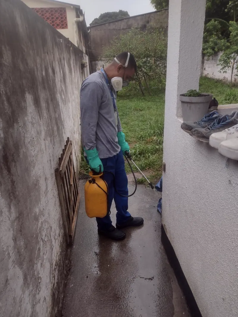 Técnico da Essencial Saúde Ambiental com pulverizador no corredor lateral de uma residência